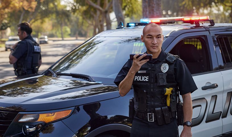 Two police officers standing outside patrol car, using FirstNet Push-to-Talk devices and FirstNet Fusion