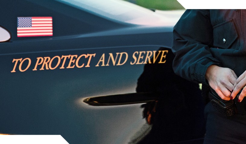 Uniformed law enforcement stands next to vehicle with a printed American flag and the slogan, "TO PROTECT AND SERVE"