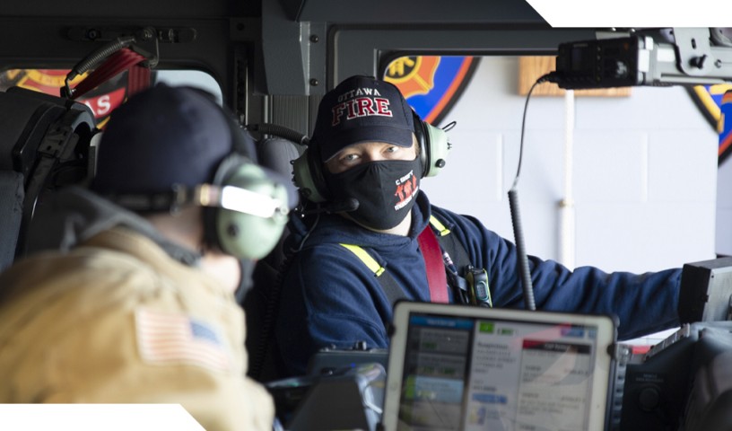 Masked Ottowan fireman sits behind the wheel with headset on, look at partner in passenger seat.